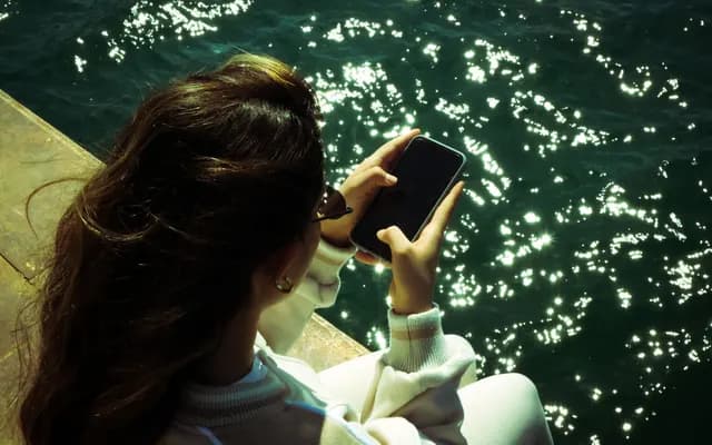 Su kenarında oturmuş, telefonuna bakan bir kadın; arkadan çekim